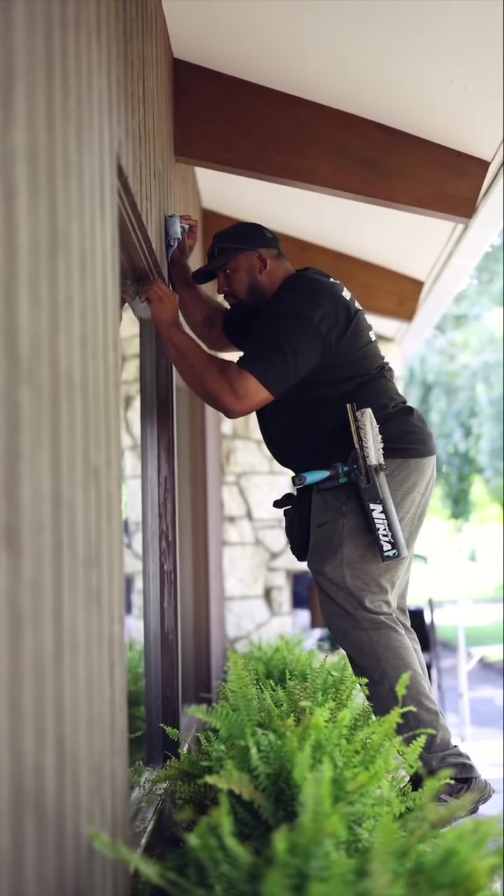 An Allen's Property Services crew member scrubbing the top of a window frame on a Yakima Valley home, ferns and stone in the foreground.